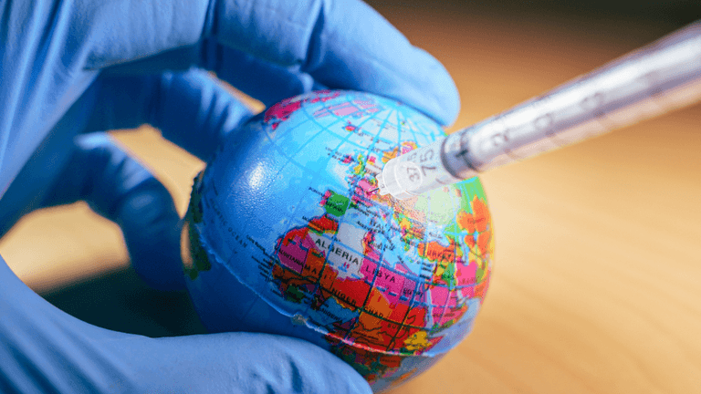 Gloved hand holding a globe while a syringe points toward it, symbolizing global vaccination.