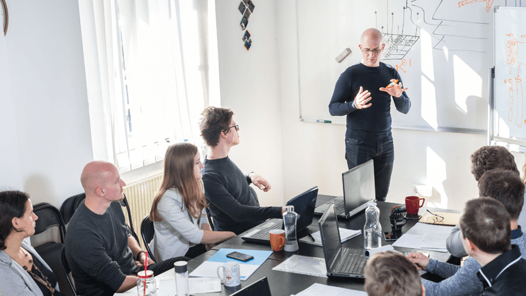 Business professional presenting to colleagues during a technology strategy meeting with laptops and a whiteboard.